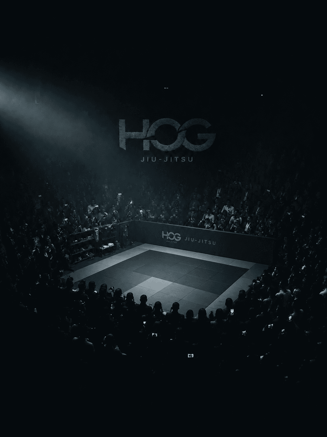 An empty HOG Jiu-Jitsu mat at center stage under a single beam of light, surrounded by a packed crowd waiting for the next match.